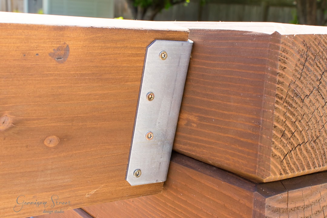 Close-up of a wooden outdoor sofa corner reinforced with a metal bracket. The bracket is fastened with screws to the wooden beams, showing clear wood grain texture and joints. Sunlight illuminates the structure.