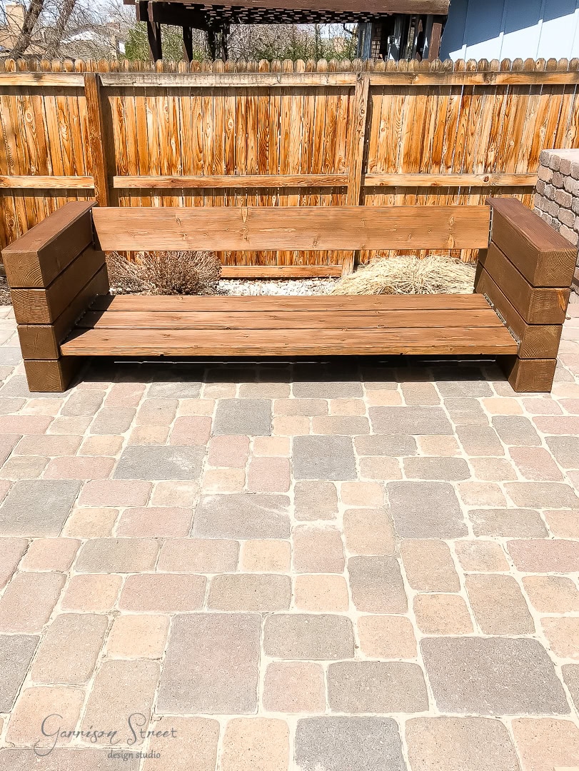 A wooden outdoor bench with a minimalistic design stands on a paved stone patio. The bench is framed by a wooden fence in the background, and there are small shrubs and a wooden pergola in the distance.