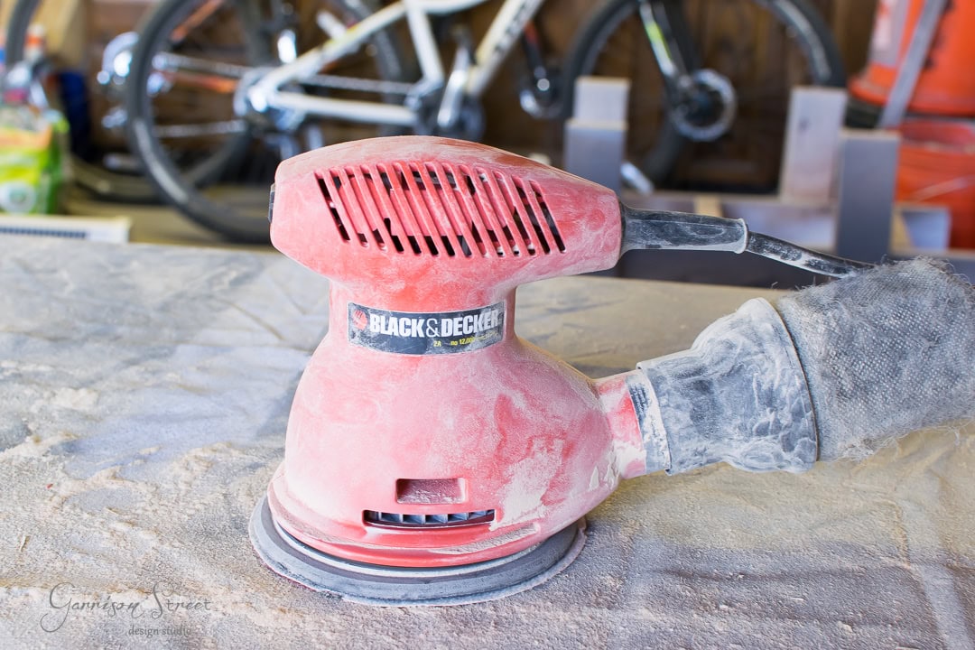 A red Black & Decker orbital sander rests on a dusty surface in a workshop. Bicycles are visible in the blurred background, suggesting a multifaceted workspace.