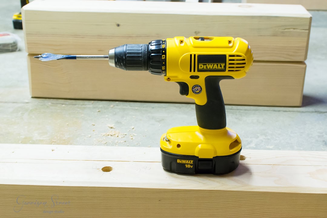 A yellow DEWALT cordless drill with an 18V battery rests on two stacked wooden planks. The drill sports a spade bit, and wood shavings are scattered nearby. In the background, more planks lie on the concrete floor, ready for the next step.