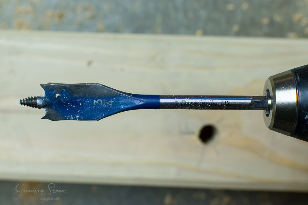 A blue spade drill bit attached to a drill is shown pointing horizontally. Behind it, a piece of wood showcases a cleanly drilled hole.