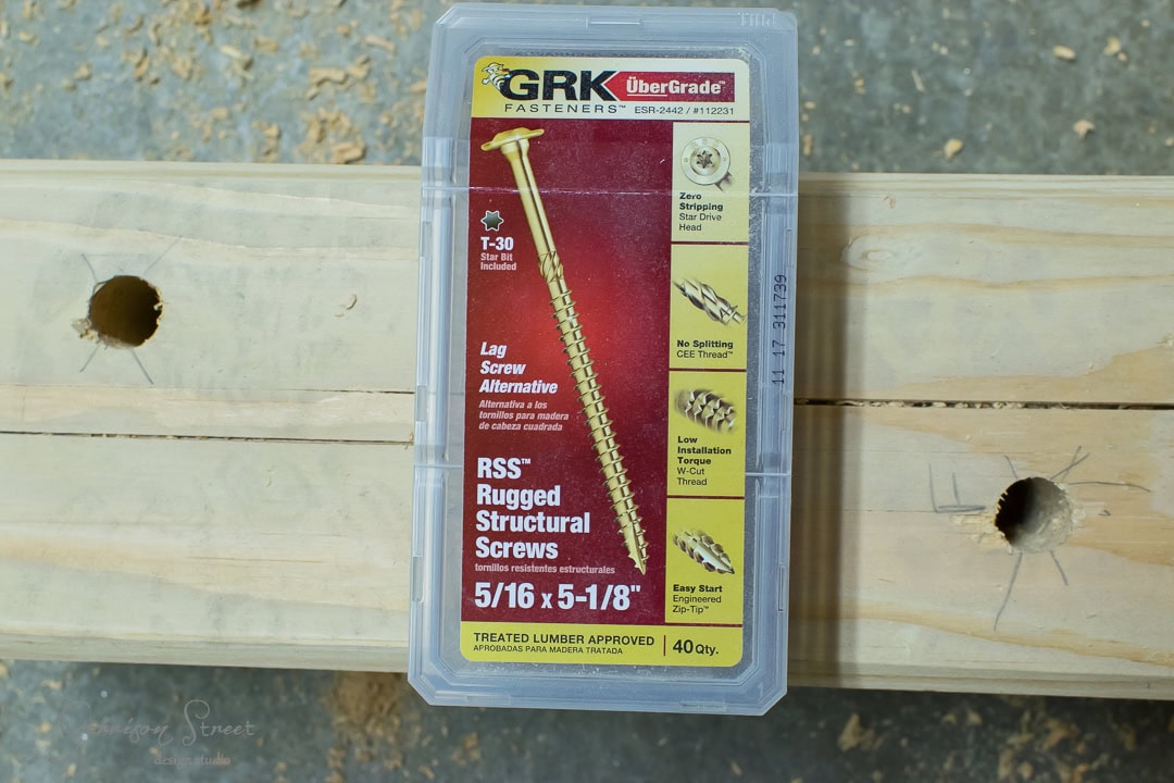 A package of GRK Fasteners UberGrade structural screws sits on a wooden surface scattered with drilled holes, perfect for your DIY outdoor sofa. The label highlights their star drive and self-tapping design, making them an excellent lag screw alternative.