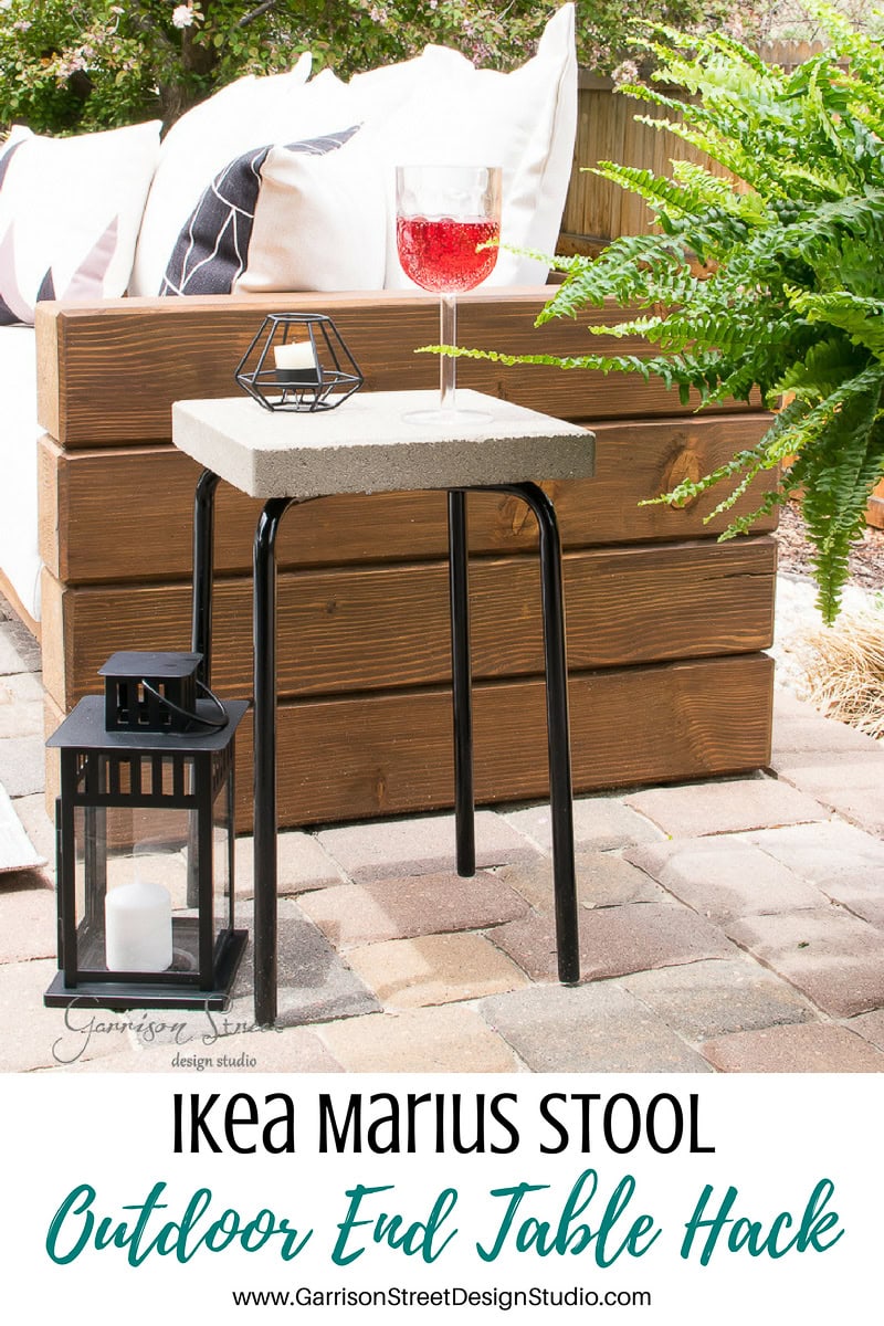 A small outdoor setup showcases an ingenious IKEA hack: the Marius stool transformed into an end table with a concrete top. It holds a glass of red beverage and a small decor item, while a black lantern rests nearby. Cushioned seating and greenery complete the inviting scene.