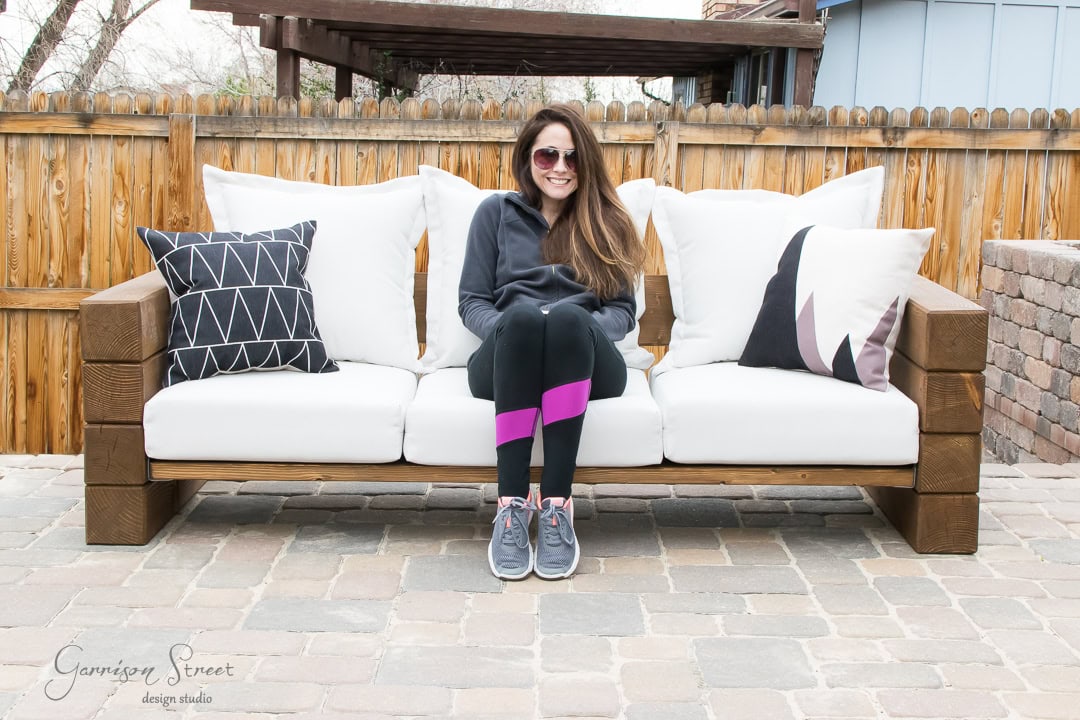 A woman wearing sunglasses and a hoodie lounges on a DIY outdoor sofa adorned with white cushions and patterned pillows. The cozy setting features a paved patio, complemented by a wooden fence and pergola in the background.