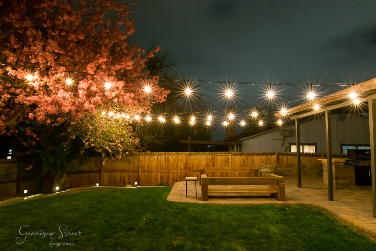 A backyard at night featuring string lights hanging above a patio area. A blooming tree with pink flowers is on the left, and there's a wooden bench on the stone patio. A wooden fence encloses the yard. The sky is overcast.