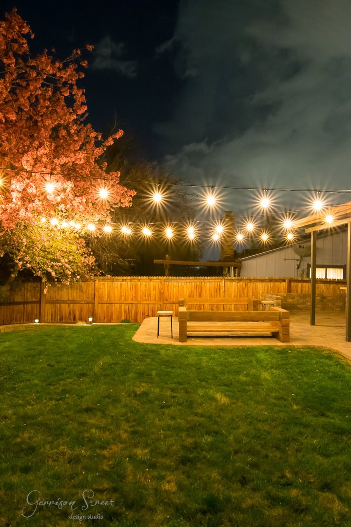 A backyard at night featuring string lights hanging above a patio with a wooden bench. A tree with pink blossoms is on the left, and a wooden fence encloses the grassy yard. The sky is cloudy.