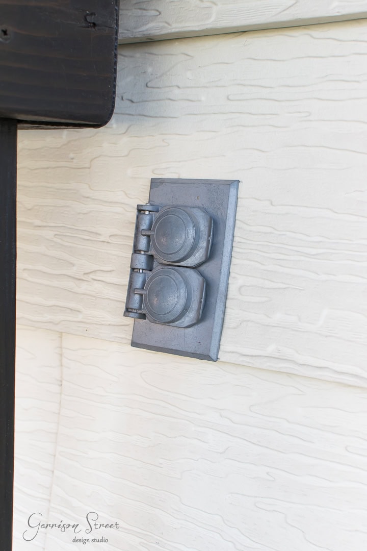 A gray metallic outdoor electrical outlet with a protective cover is mounted on cream-colored siding. The siding has a textured, wood-like appearance.