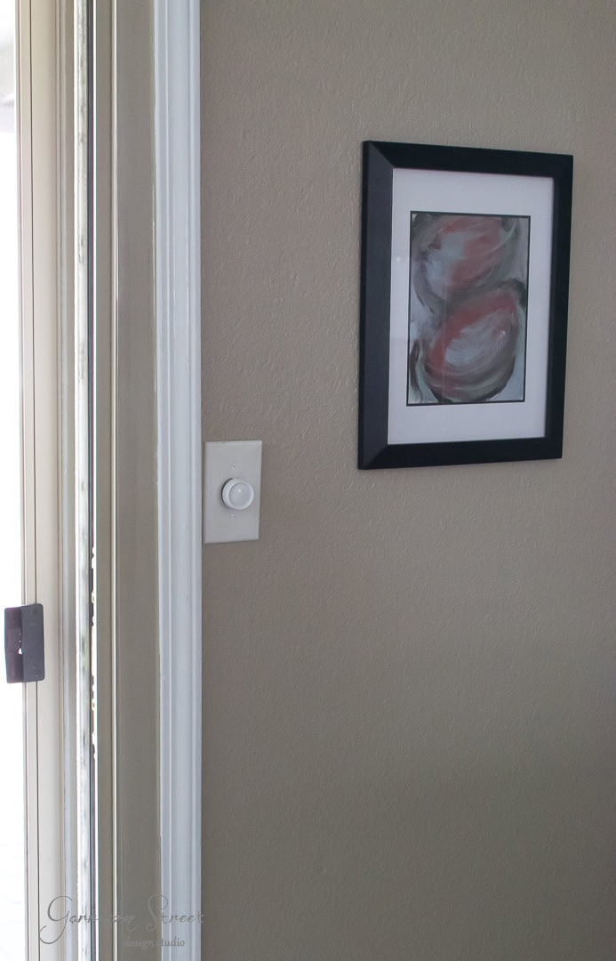 A beige wall features a framed abstract painting with swirls of black, white, and red. A round thermostat is mounted nearby. The edge of a glass door is visible on the left.