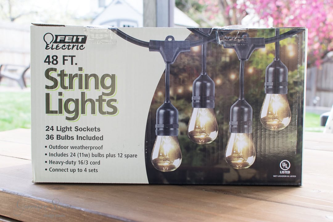 A box of Feit Electric 48 ft. outdoor weatherproof string lights with 24 light sockets and 36 bulbs, including 12 spare bulbs. The package is on a wooden table with a window and blooming trees in the background.