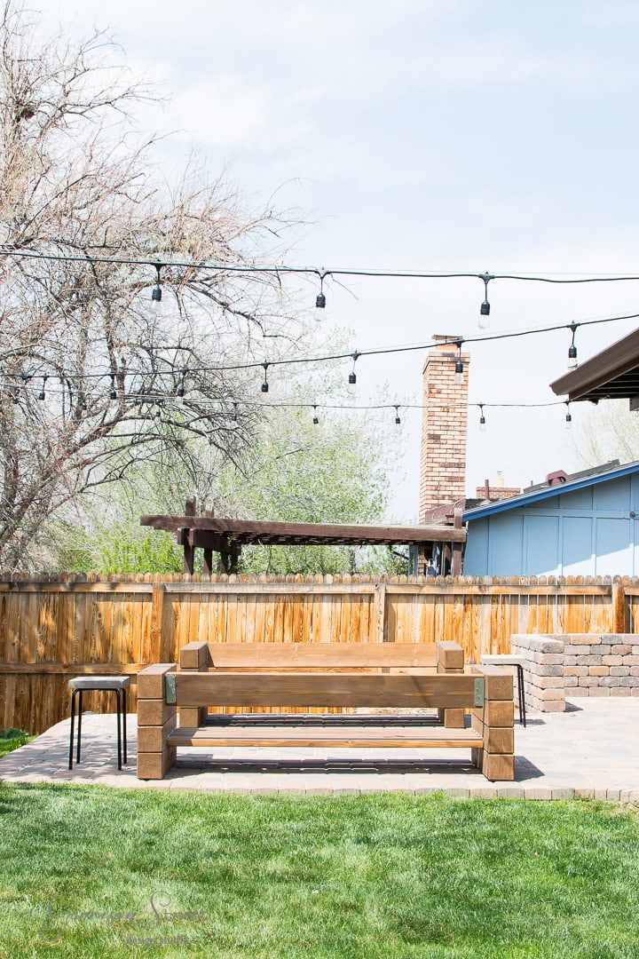 A backyard with a wooden fence, string lights overhead, and a wooden bench on a paved patio. Leafless trees and a chimney are visible in the background under a clear sky.