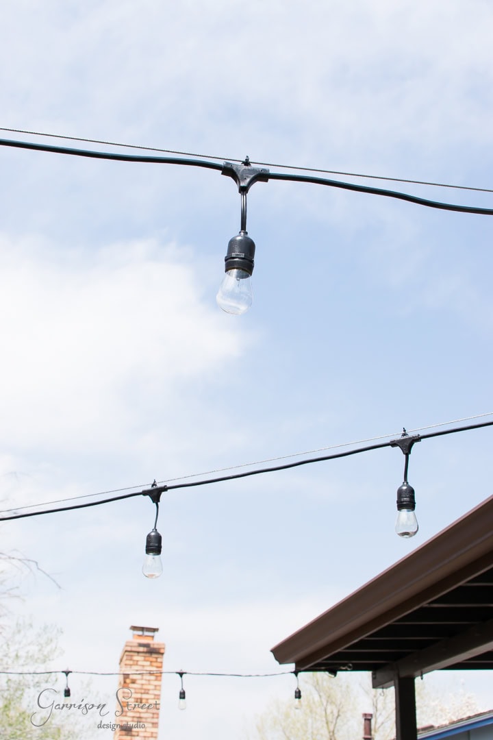 String lights hang across an outdoor space against a clear blue sky. A chimney and part of a roof are visible in the background.