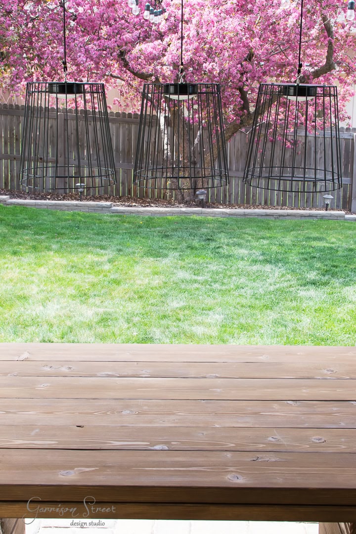 A wooden outdoor table sits in the foreground, overlooking a vibrant green lawn. Three black wire pendant lights hang above, and a garden with blooming pink cherry blossom trees and a wooden fence are in the background.