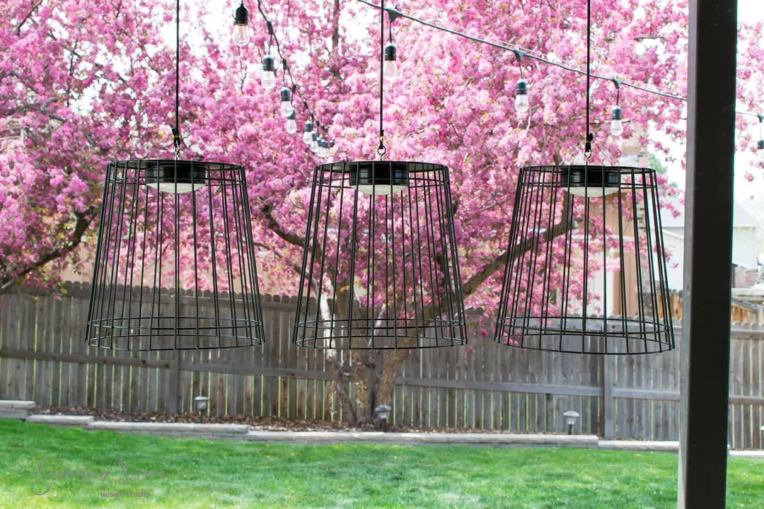 Three wire-frame pendant lights hang in front of a blossoming cherry tree with pink flowers. A wooden fence and a green lawn are visible in the background.