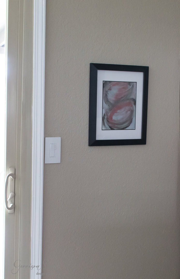 A framed abstract painting with pink and gray shades hangs on a beige wall near a sliding glass door and a light switch.