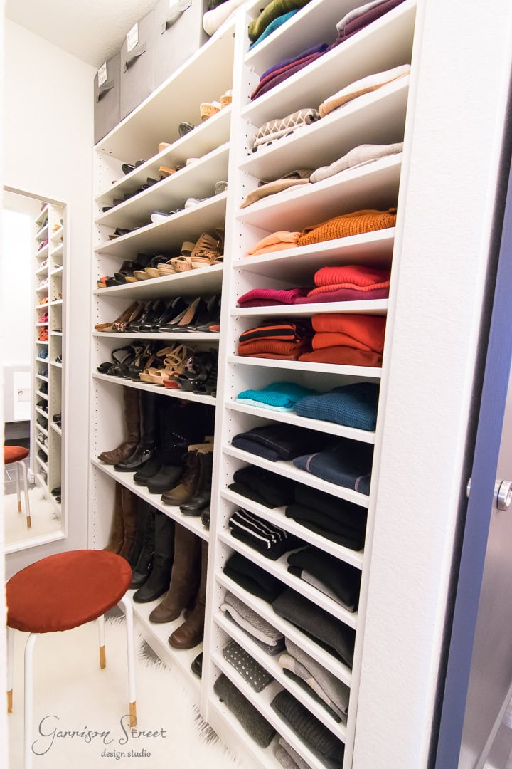A neatly organized walk-in closet boasts a thoughtful design with shelves filled with folded sweaters, shoes, and boots. A red cushioned stool rests on a white rug in front of the shelves, while a mirror is mounted on the left wall.