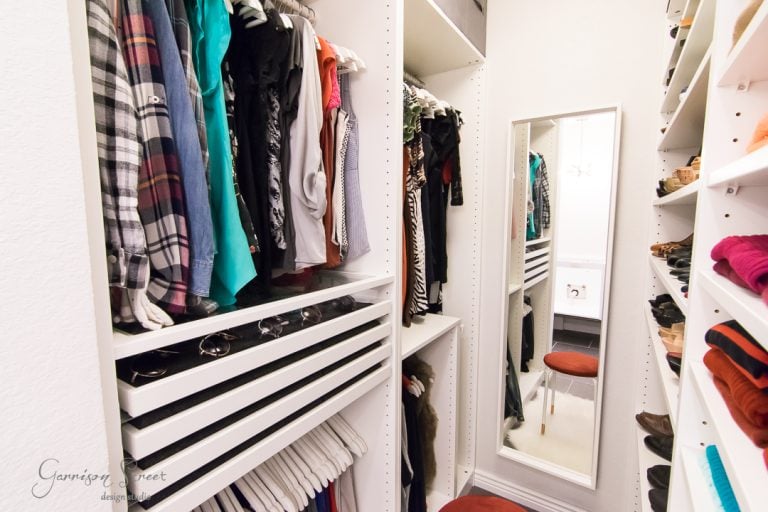 A neatly organized walk-in closet with white shelving. Clothes are hung on racks, and shoes are arranged on side shelves. A long mirror is mounted on one wall, and a small red stool is placed in front of it.