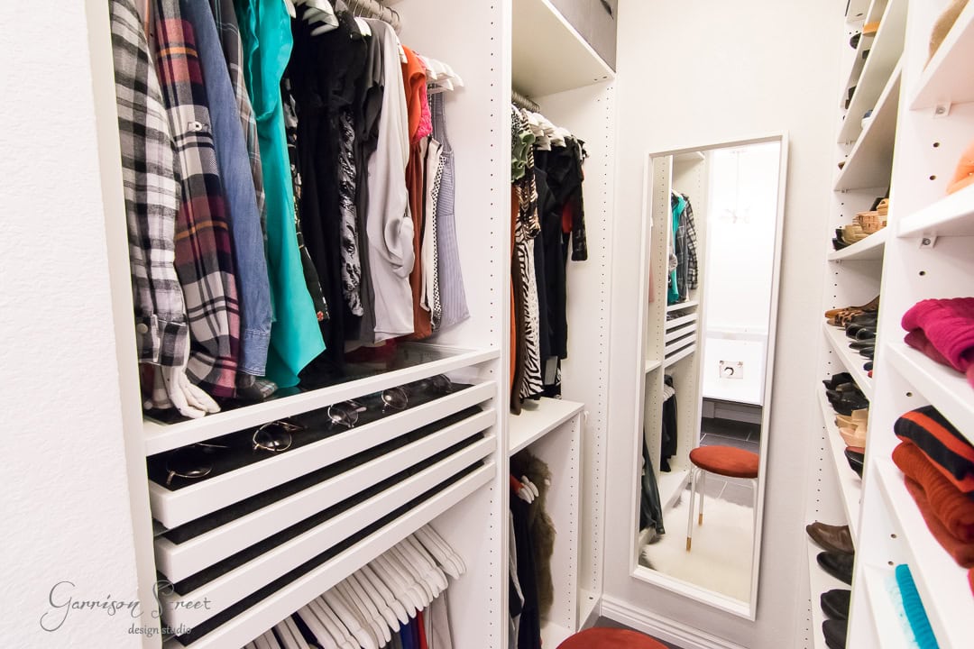 A neatly organized walk-in closet with white shelving. Clothes are hung on racks, and shoes are arranged on side shelves. A long mirror is mounted on one wall, and a small red stool is placed in front of it.