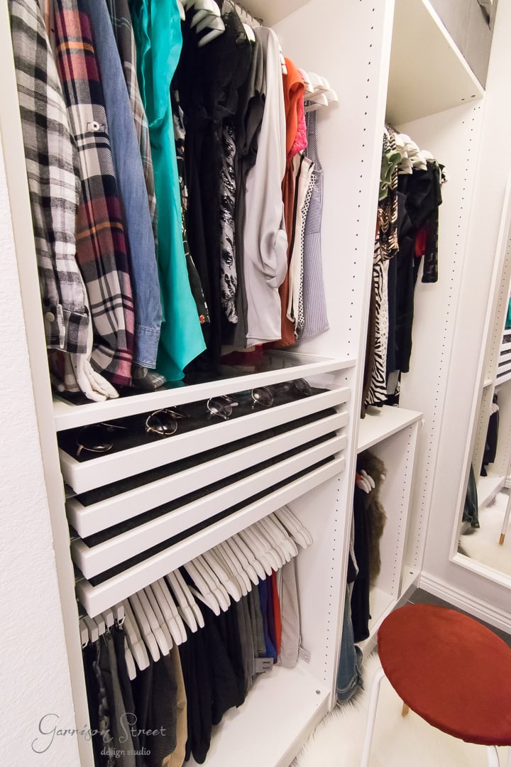 A neatly organized closet design features folded shirts on shelves and clothes hanging below. A mirror leans against the wall, while a small red stool perches on a white rug in the foreground.