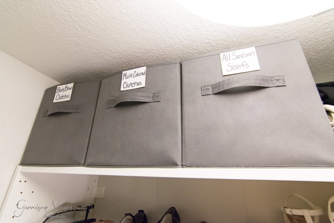 Three gray storage boxes labeled "Black/Brown Clutches," "Multi-Colored Clutches," and "All Season Scarfs" sit neatly designed on a white closet shelf, with a row of shoes underneath.