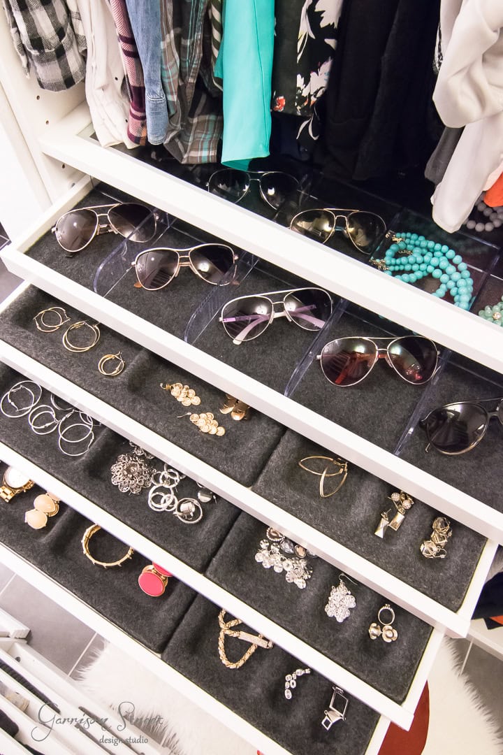 A well-organized jewelry drawer, seamlessly integrated into the closet design, features four levels. Each level showcases accessories like sunglasses, rings, earrings, bracelets, and necklaces. The drawer has a white frame with black velvet-like interiors for elegant display.