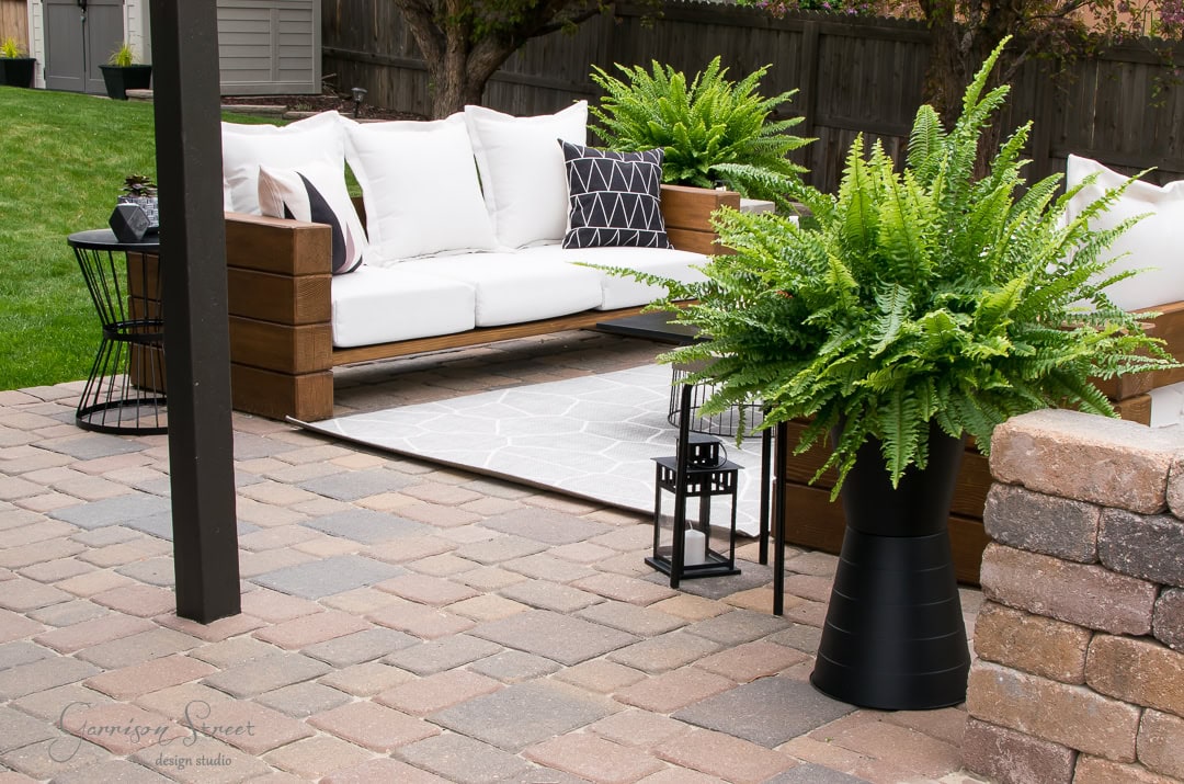 Modern outdoor patio with a wooden sofa featuring white cushions and patterned throw pillows. Large ferns in sleek black planters decorate the space. A black side table and lantern accent the stone-paved floor beside a gray rug, adding a stylish touch.