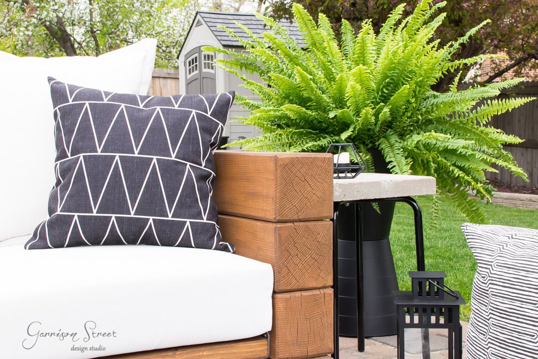 A cozy outdoor patio setup featuring a modern wooden chair with white cushions and a black geometric-patterned pillow. An IKEA fniss adds storage next to lush green ferns and decorative lanterns, enhancing the tranquil garden ambiance. A small gray shed is visible in the background.
