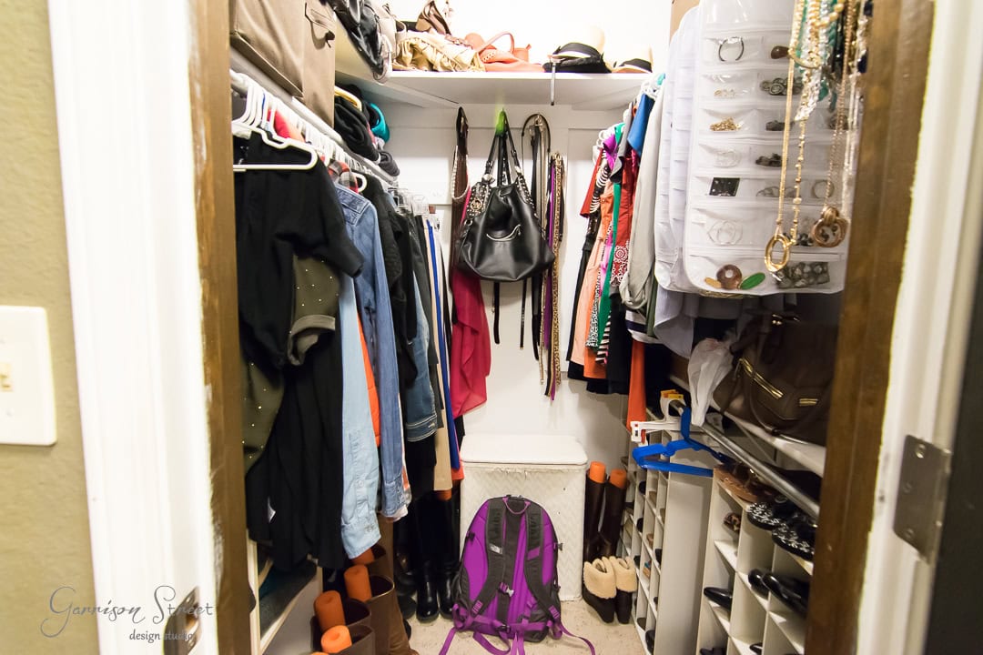 A small, neatly designed closet brims with clothes hanging on rods and accessories like hats, bags, and shoes. A purple backpack rests on the floor while a shoe organizer graces the door. Shelves showcase a thoughtful design with neatly arranged purses and folded items.