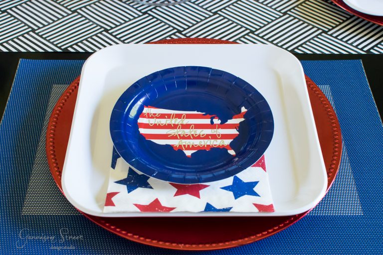 A patriotic tablescape features a blue USA-shaped paper plate on a white square plate, adorned with a quote in white and red. A star-patterned napkin rests beneath, atop a red charger. The backdrop is an engaging geometric pattern.