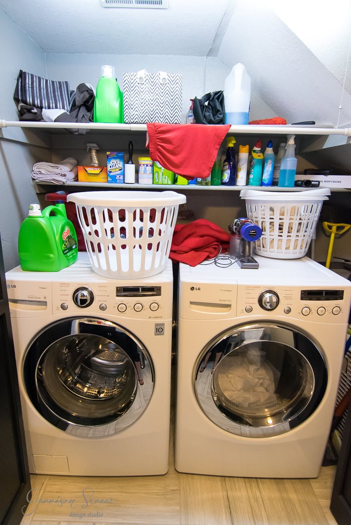 A bright and organized laundry room features a front-loading washer and dryer with two baskets perched above. On the shelf, cleaning supplies, detergents, and neatly folded clothes are perfectly arranged, showcasing efficient organization.