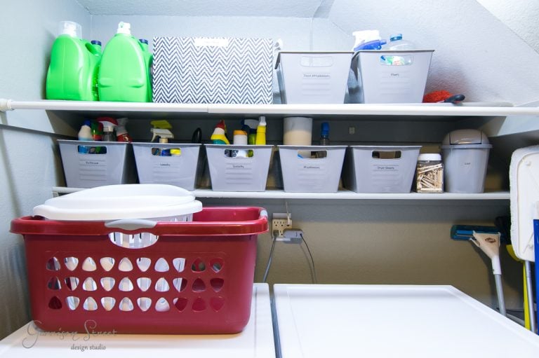 A laundry room with organized shelves. White plastic bins labeled for various cleaning supplies sit on the shelves. Two large green detergent bottles are visible. A red laundry basket is on the washing machine below. A mop is in the corner.