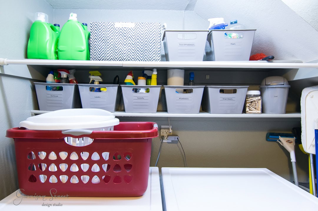 A laundry room with organized shelves. White plastic bins labeled for various cleaning supplies sit on the shelves. Two large green detergent bottles are visible. A red laundry basket is on the washing machine below. A mop is in the corner.