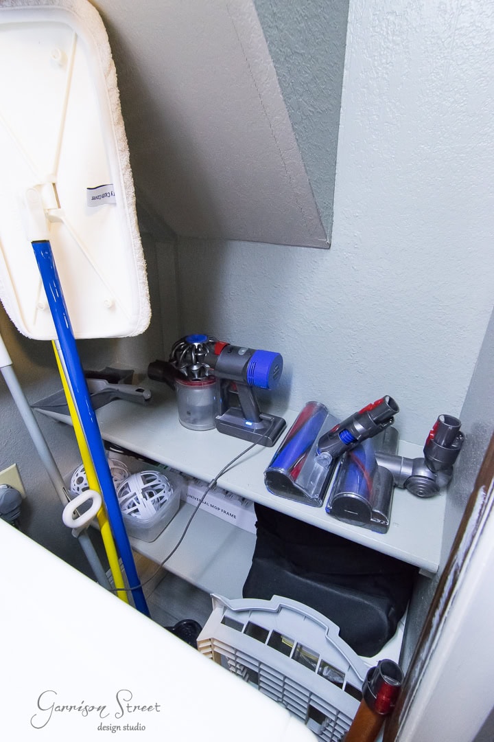 A small, organized closet storage space contains a vacuum cleaner with various attachments on a shelf, an ironing board, a mop, and other miscellaneous cleaning supplies. Perfect for keeping your laundry room tidy, the wall is painted light gray.