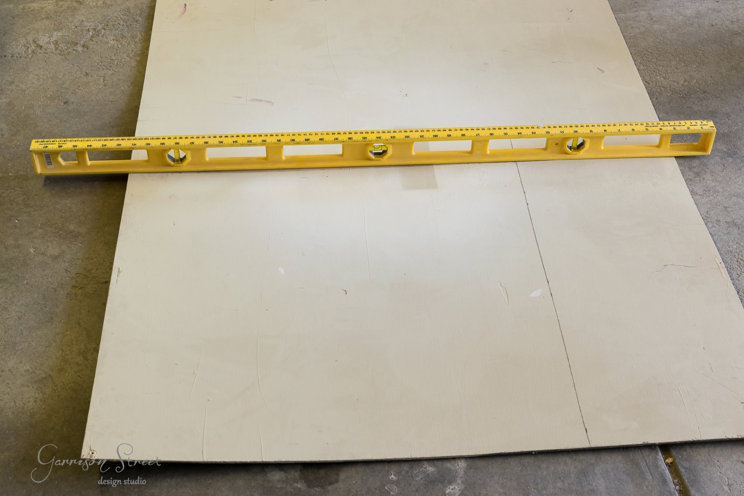 A yellow spirit level rests on a large sheet of drywall lying on a gray concrete floor, like logs ready to be stacked. The level, with its trio of bubble vials, ensures precision for any task—even the perfect installation of a cozy fireplace insert.