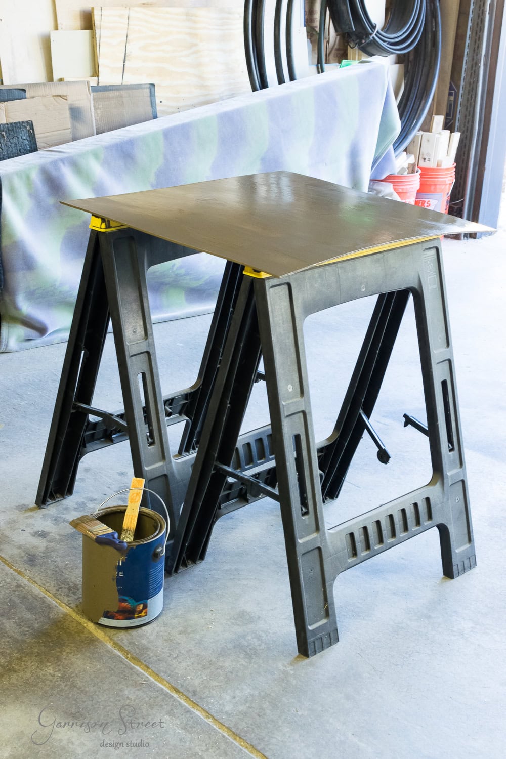 A pair of black sawhorses with a wooden board on top stand in a workshop resembling logs stacked by a fireplace insert. Nearby, there's a bucket containing paintbrushes and a can of paint. Rolls of material and red buckets are visible in the background.