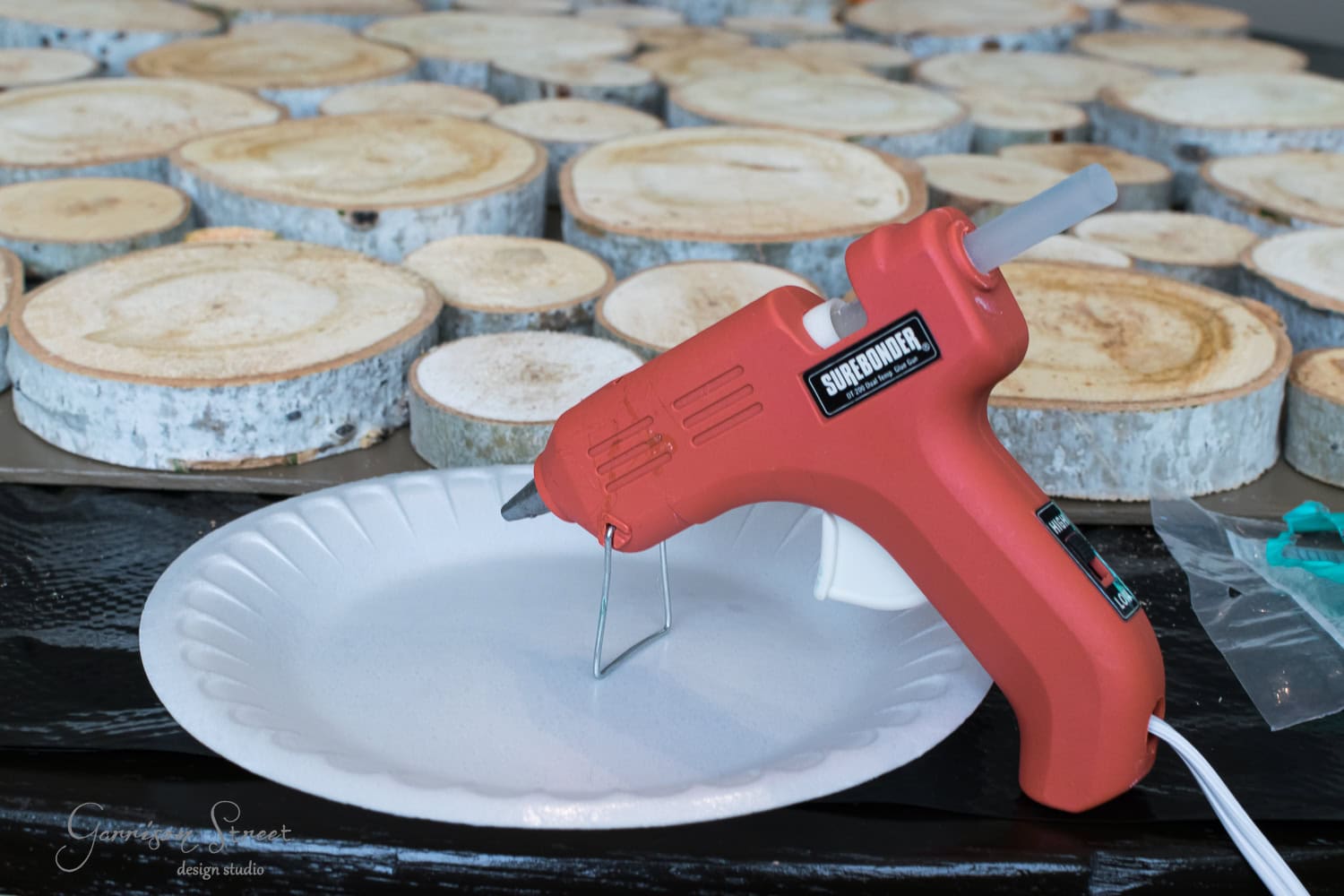 A red hot glue gun rests on a white paper plate, surrounded by various wooden slices resembling mini logs on a black surface, ready for a craft project.
