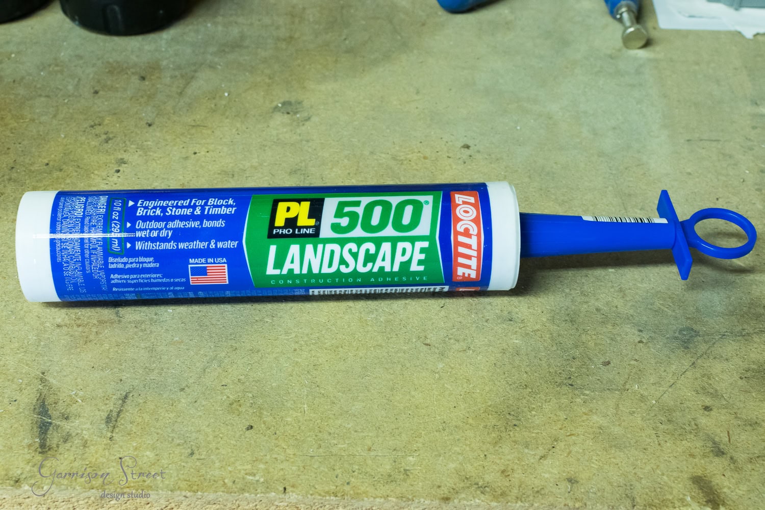 A tube of Loctite PL 500 Landscape Construction Adhesive rests on a workbench, ready to secure everything from stones to logs. The tube is blue and white with a green and yellow label featuring a push nozzle. Tools and other workshop items are visible in the background.