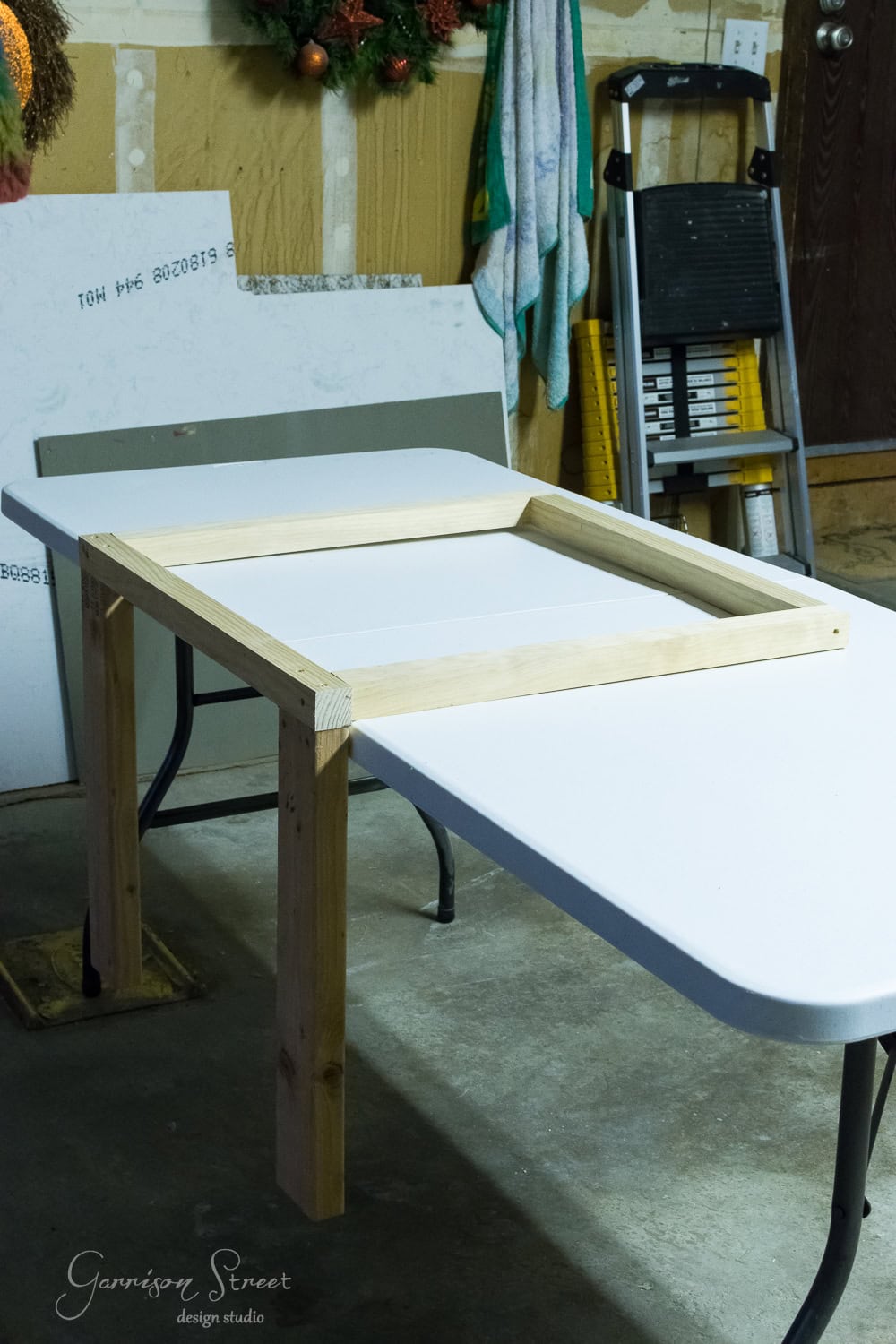 A wooden frame is partially assembled on a white folding table in a garage. A ladder and other tools are visible in the background, next to a fireplace insert awaiting installation, with a piece of drywall resting against the wall.