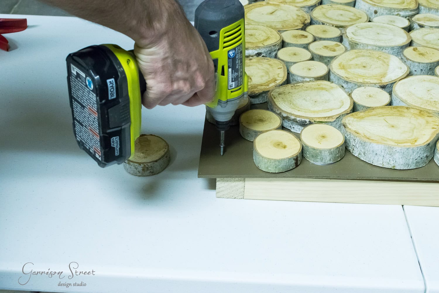 A person uses a yellow power drill to attach a wooden slab to a base covered with circular wood pieces cut from tree branches, resembling miniature logs. The surface is on a white table.