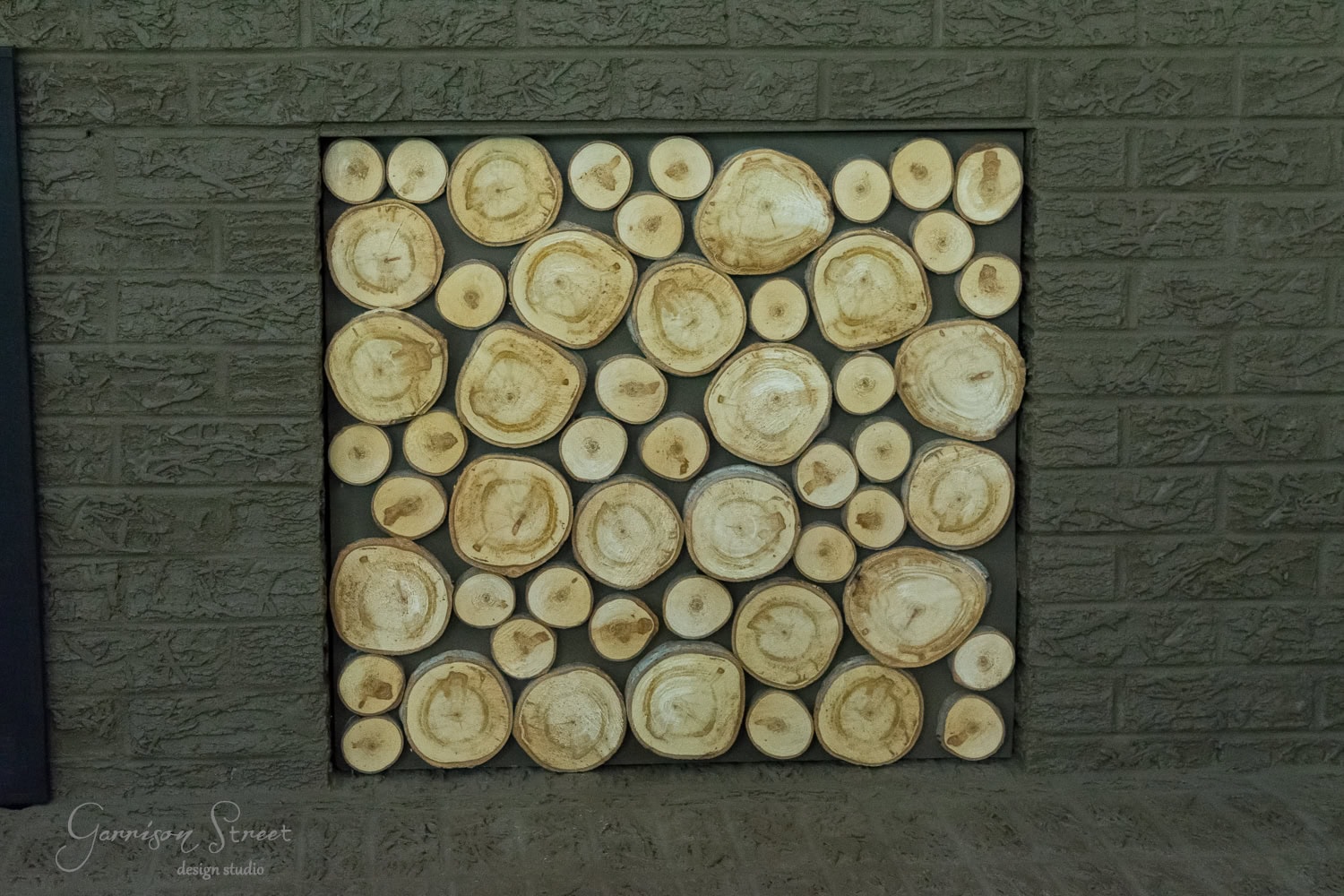 A decorative arrangement of circular wood slices fills a square opening in a brick wall, resembling a rustic fireplace insert and creating a natural display with the charm of stacked logs.