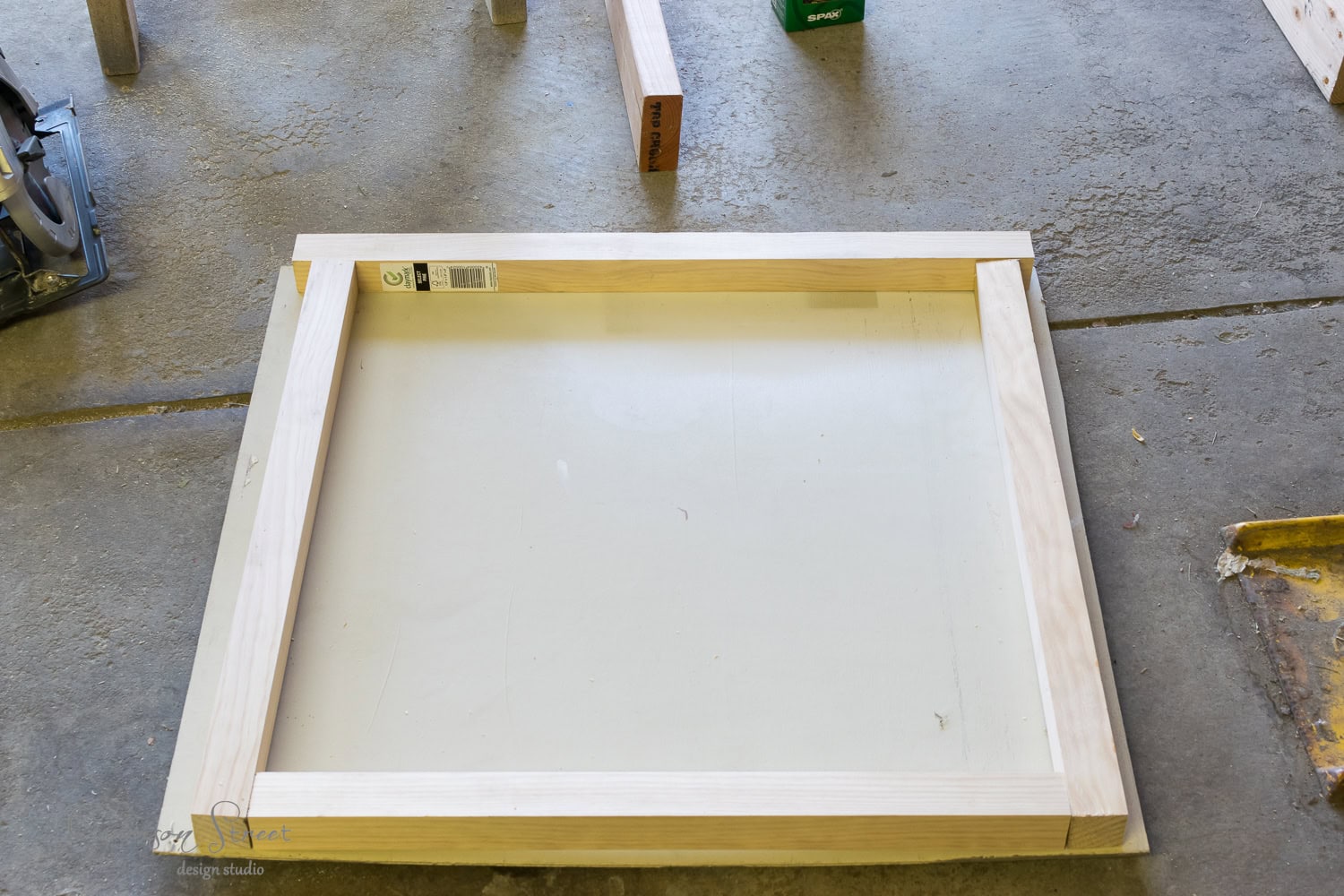 A partially assembled wooden frame lays on a concrete floor, surrounded by tools, wooden pieces, and a stack of logs ready to feed the nearby fireplace insert.