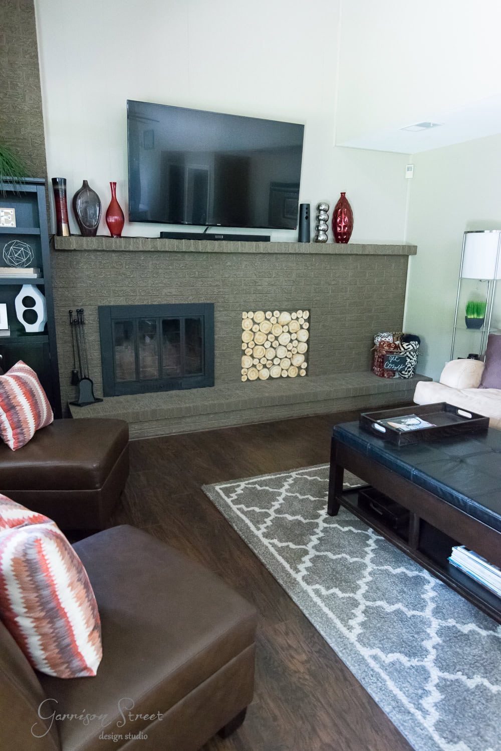 A cozy living room features a flat-screen TV mounted above a brick fireplace, enhanced by a modern fireplace insert. The mantel displays red and black vases, while two brown armchairs with striped pillows surround a dark wood coffee table resting on a patterned rug.