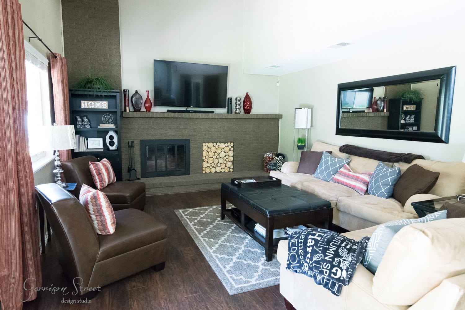 A cozy living room features a large beige sectional sofa, two brown armchairs, and a dark coffee table. A TV is mounted above the fireplace insert with wood decor, creating warmth. Shelves with decor and logs, along with a large mirror, elegantly adorn the walls.