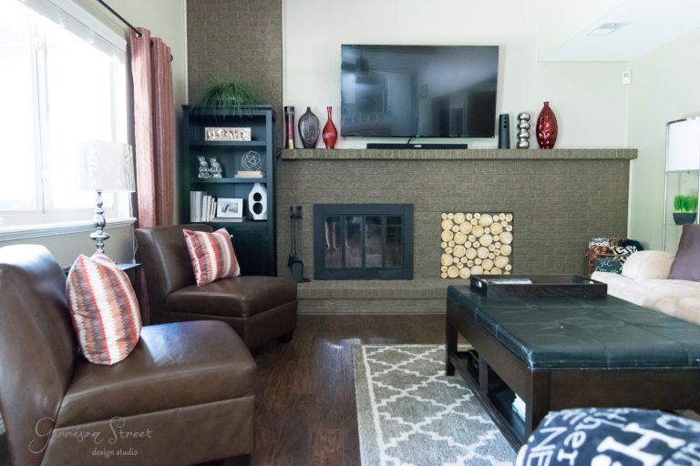 A cozy living room with a large TV above a fireplace, decorated with vases and art pieces. Two brown leather chairs, a patterned rug, and a black coffee table add to the earthy tones. A stack of firewood fills the hearth below.