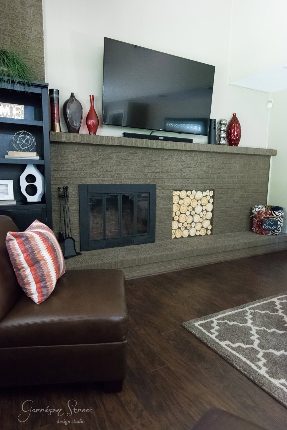 In the living room, a flat-screen TV sits above a brick fireplace boasting a modern fireplace insert. Decor includes vibrant red vases, a wooden log panel, and decorative balls. A brown sectional sofa with patterned pillows rests on dark wooden flooring beside a geometric-patterned rug.