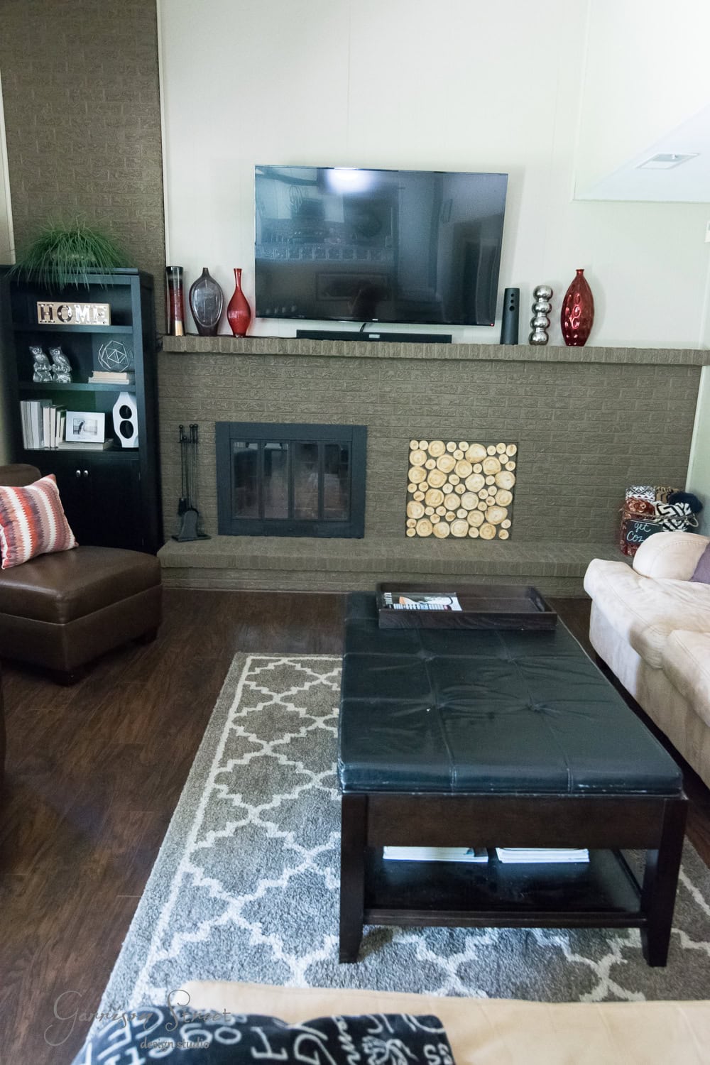 A cozy living room showcases a large TV mounted above a brick fireplace insert. The space features a dark wood coffee table on a patterned rug, brown and beige seating, decorative vases, and a neatly stacked pile of logs by the inviting fireplace.