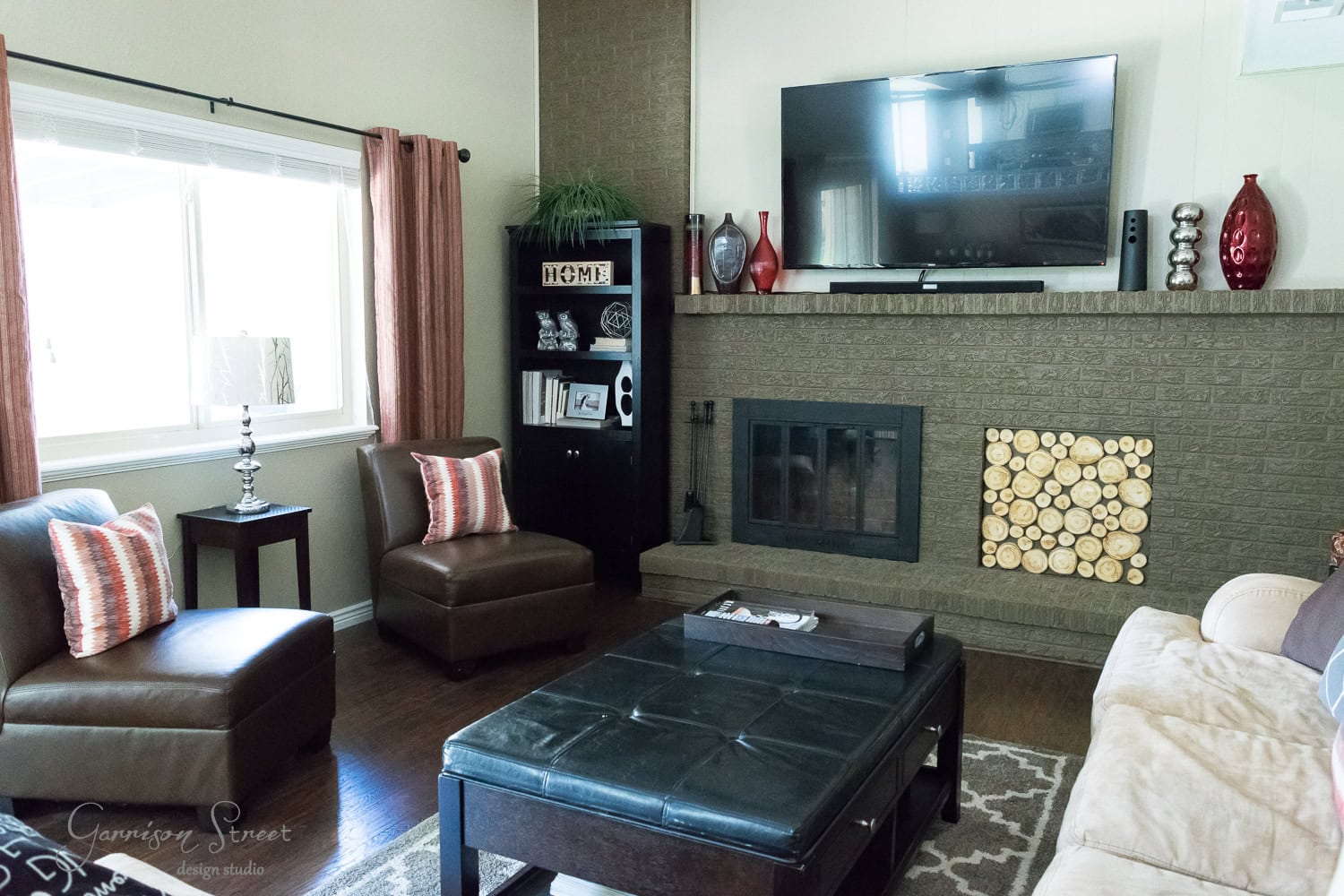 A cozy living room features a large dark leather ottoman, two brown armchairs with striped cushions, and a fireplace insert with a stylish wood stack design. A flat-screen TV is mounted above the fireplace, and a window with red curtains brightens the space.