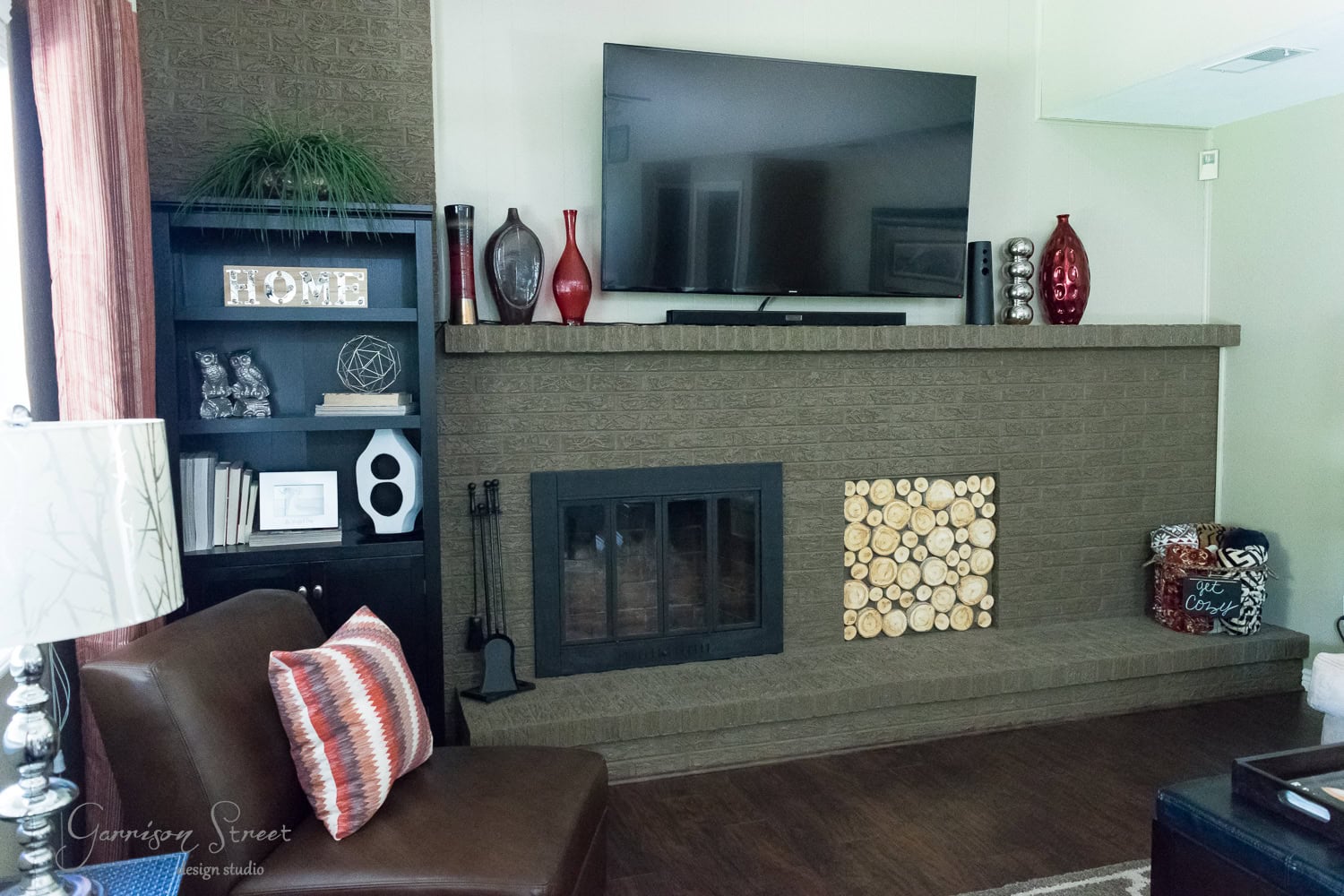 A cozy living room features a large brick fireplace insert with a mounted TV. The mantel is decorated with candles and vases, while neatly stacked logs are arranged beside the hearth. A comfy chair with a colorful cushion sits nearby, perfect for unwinding.