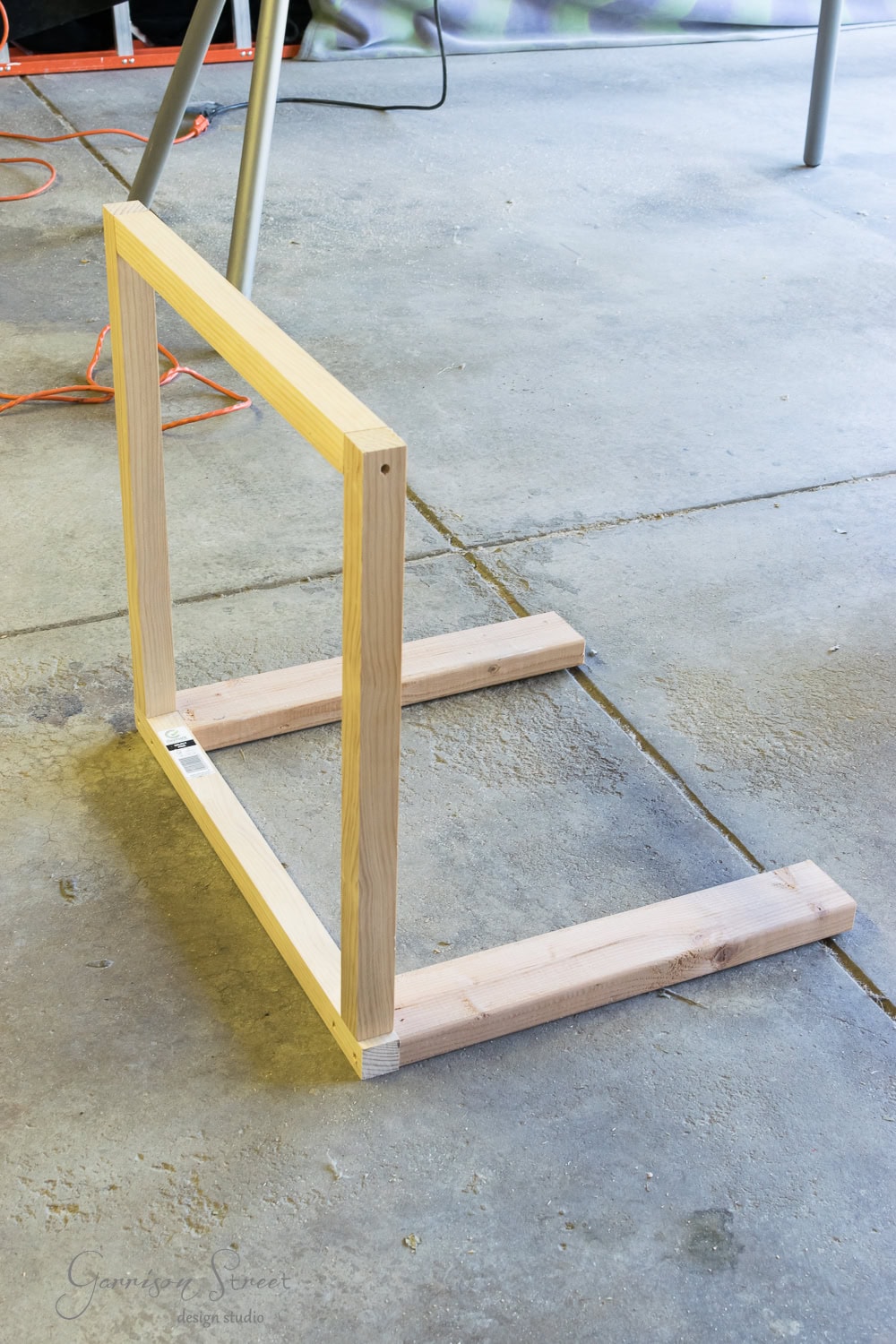 A wooden frame structure is being assembled on a concrete floor, resembling the framework for a fireplace insert. It consists of two vertical beams connected by horizontal ones at the bottom, forming a rectangular shape, with tools and cords scattered around like logs waiting to be organized.