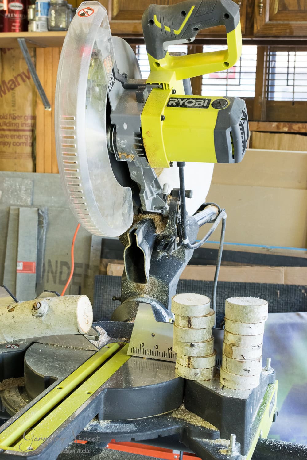 A green and black Ryobi miter saw is shown in a workshop, surrounded by tools. A pile of cut wooden discs is stacked on the saw table, with a fresh cut log—perfect for a fireplace insert—placed to its left. Tools and materials fill the background.