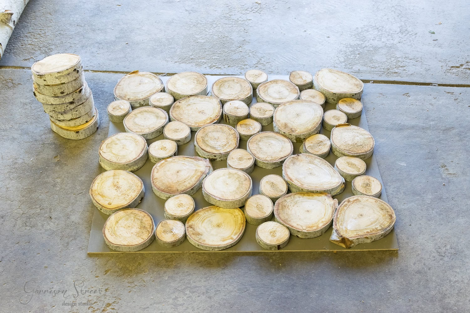 A collection of round wood slices, reminiscent of logs for a fireplace insert, is arranged on a metal sheet on a concrete floor. A stack of similar wood slices rests nearby on the left.